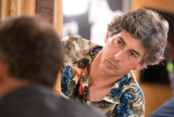 A man with wavy gray hair wearing a blue and red floral shirt looks forward with a serious expression.