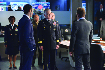 Military officers in dress uniforms and men in suits stand in a high-tech command center.