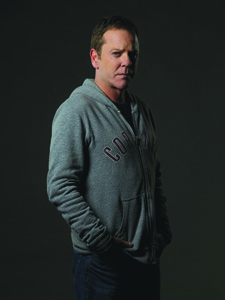 A man wearing a grey zip-up hoodie and dark pants stands against a dark background.