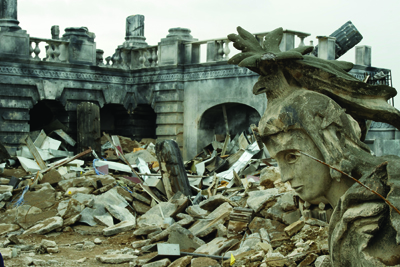 A weathered stone statue lies partially buried in a pile of concrete debris and rubble in front of a ruined building.