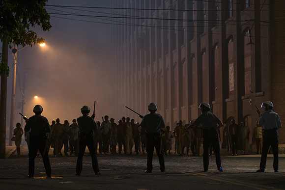 Silhouetted security officers stand in a line facing a crowd in a smoky street at night.