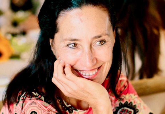 A smiling woman with dark hair rests her chin on her hand while looking off-camera.