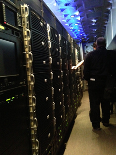 A person walks down a narrow aisle lined with tall, black server racks under blue ceiling lights.
