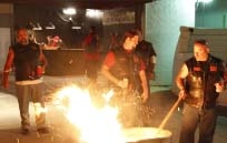 Several men in dark vests stand around a large, bright bonfire at night.