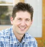 A man with short dark hair smiling while wearing a blue and white plaid button-down shirt.