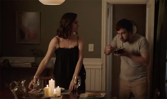 A woman in a black slip dress looks toward a man eating from a bowl in a doorway.