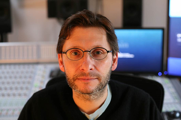 A man with short brown hair and glasses sits in front of a mixing console and computer monitors.