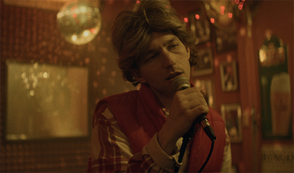 A man in a red vest holds a microphone to his mouth in a dimly lit room with disco lights.