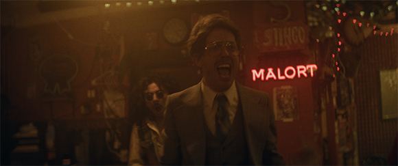 A man in a suit and glasses laughs with his mouth wide open in a dimly lit, cluttered room.