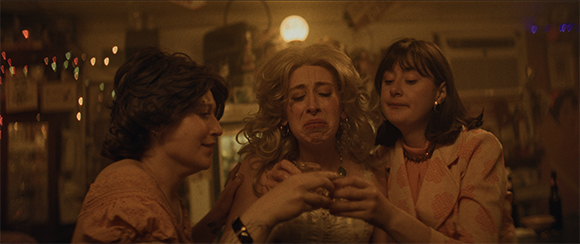 Three women in a warm-toned room; the center woman is crying while the others hold her hands.