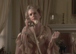 A blonde woman in a pink ruffled dress talks on a gold vintage telephone in a dimly lit room.