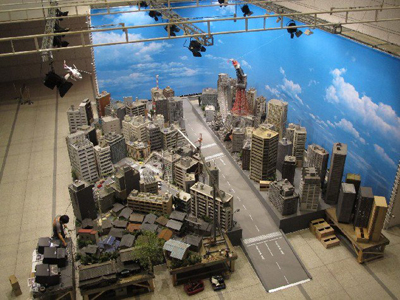 A miniature cityscape model with buildings and a road set against a large blue sky backdrop in a studio.