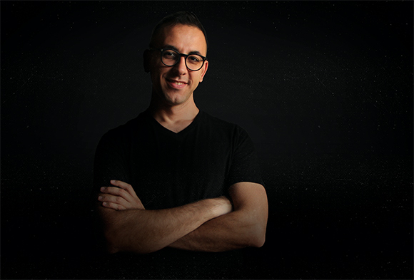 A smiling man wearing glasses and a black t-shirt stands with his arms crossed against a dark, speckled background.
