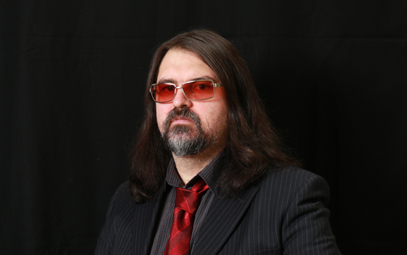 A man with long dark hair, a goatee, and red-tinted glasses wearing a dark pinstripe suit and red tie.