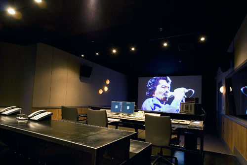 A dark sound mixing studio featuring a large desk, audio equipment, and a projection of a man singing.