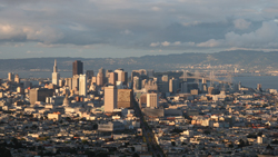 Aerial view of the San Francisco skyline with skyscrapers and a bridge under a cloudy sky.