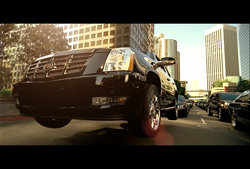 Low-angle view of a black SUV driving down a sunlit city street lined with skyscrapers.