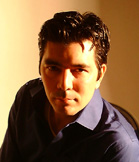 A man with dark hair and a blue button-down shirt looks toward the camera under warm lighting.