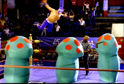 A wrestler in blue jumps through the air above three large, blue, mushroom-shaped costumes in a wrestling ring.