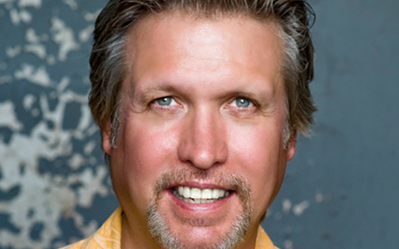 Close-up portrait of a smiling man with light brown hair, blue eyes, and a groomed goatee.