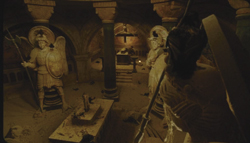 Two large angel statues with wings and spears stand in a dark, vaulted stone room.