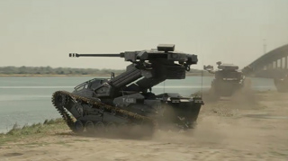 A dark, heavy military tank with a long cannon drives through dust near a body of water.