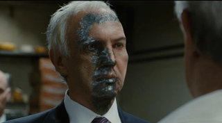 A man with dark, textured residue covering half of his face looks toward another person.