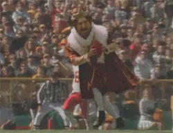 A person in a crown and red royal robe runs across a football field in front of a crowd.