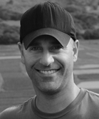 A smiling man wearing a dark baseball cap and a t-shirt.