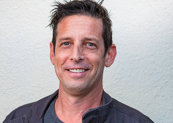 A smiling man with short dark hair looking toward the camera against a light textured wall.