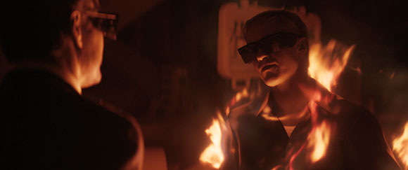 A man wearing dark sunglasses stands in a dark setting with flames rising around his torso.