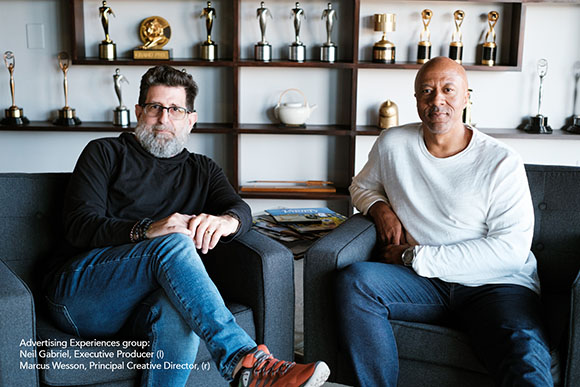 Two men sit in grey armchairs in an office with shelves of awards in the background.