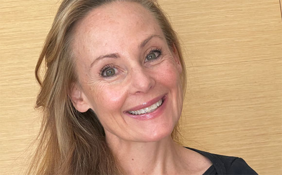 A smiling woman with blonde hair and light-colored eyes looking towards the camera.