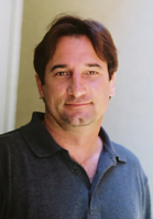 A man with brown hair and a goatee wearing a dark polo shirt, smiling against a neutral background.