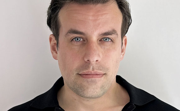 A close-up headshot of a man with dark hair and blue eyes against a plain white background.
