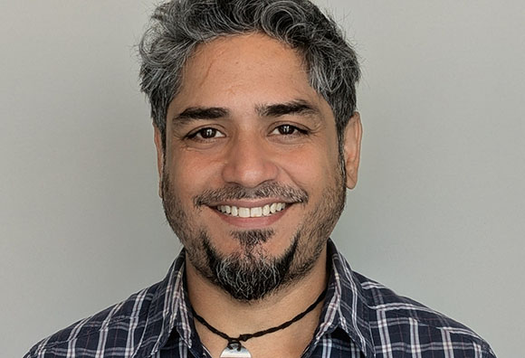 A smiling man with wavy grey hair, a beard, and a plaid shirt against a plain background.