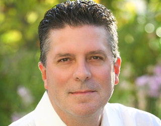 A man with dark, styled hair and a white collared shirt looks forward against a blurred green background.