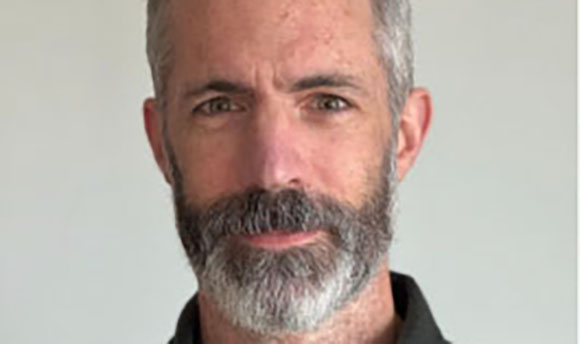 A close-up portrait of a man with graying hair and a salt-and-pepper beard against a white background.