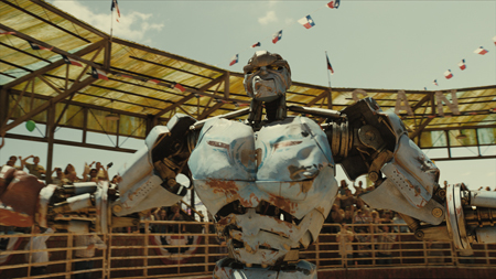 A large, weathered blue and silver robot stands in a crowded outdoor rodeo arena under a sunny sky.