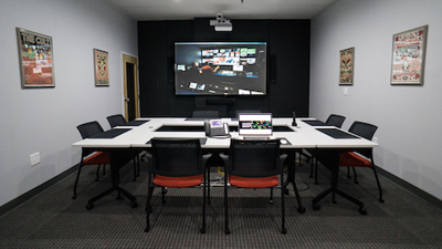 A conference room featuring a large wall-mounted monitor, a white U-shaped table, and several black and orange chairs.