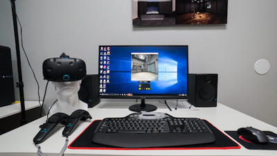 A computer monitor, keyboard, mouse, VR headset on a mannequin head, and speakers on a white desk.