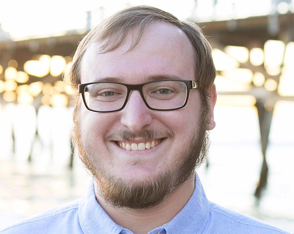 A smiling man with brown hair, glasses, and a beard wearing a blue shirt against a blurred pier background.