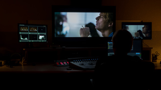 A person sits in a dark room facing multiple monitors displaying video footage and editing tools.