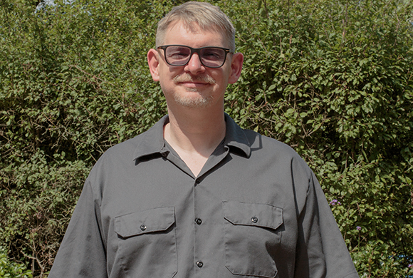 A man with glasses and a grey button-down shirt stands before a backdrop of dense green foliage.