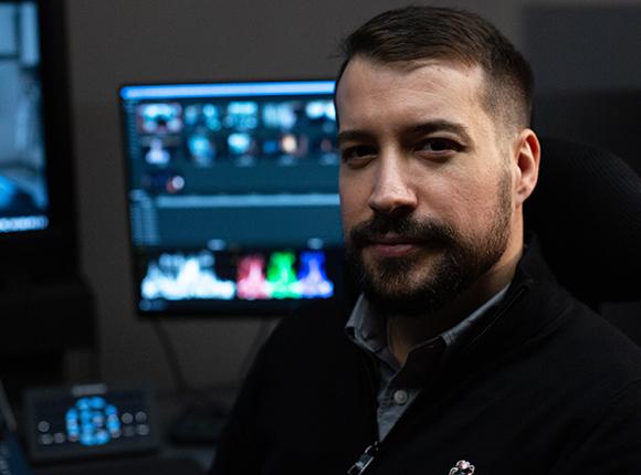 A man with a beard and short dark hair looks toward the camera in a dimly lit workspace with monitors.