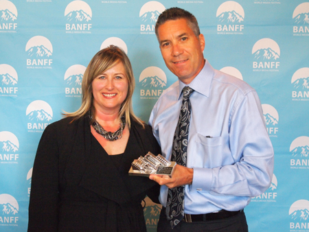 A man in a blue shirt and tie and a woman in a black dress hold a small award against a blue festival backdrop.