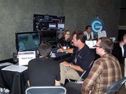 Three men and a woman stand and sit around a table with computers and displays at a conference booth.