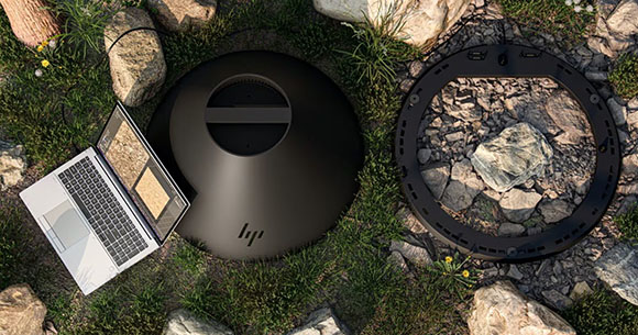 A laptop and a large black circular HP device sit on grass and rocks next to a black ring.