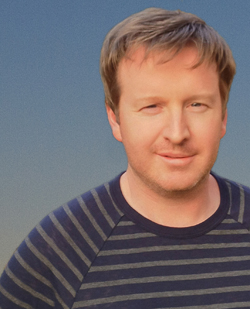 A man with light brown hair and a slight smile wearing a navy and grey striped shirt.