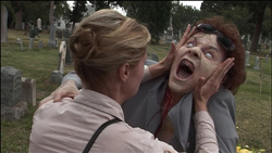 A woman grips the face of a person with pale skin and wide eyes who is screaming in a graveyard.